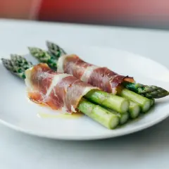 Dwa pęczki zielonych szparagów owinięte w cienkie plastry szynki parmeńskiej, polane oliwą, na białym talerzu.