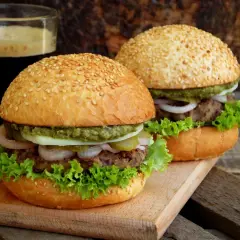 Dwa domowe burgery z sezamem, sałatą, cebulą i sosem, obok szklanki ciemnego napoju.