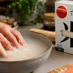 Ręce płuczą ryż do sushi w misce, przygotowując go do gotowania. Obok pudełko ryżu i łosoś.