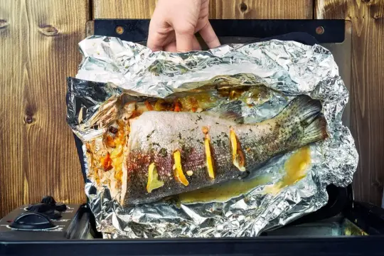 Ręka wyjmuje z piekarnika rybę z piekarnika, pieczoną w folii aluminiowej z cytryną i ziołami.