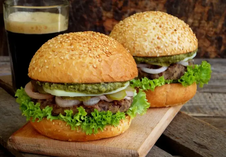 Dwa domowe burgery z sezamem, sałatą, cebulą i sosem, obok szklanki ciemnego napoju.