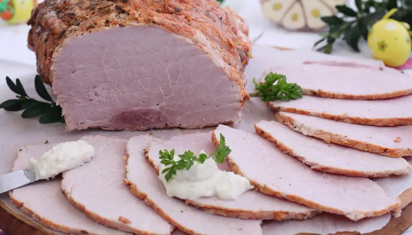 Soczysta szynka pieczona w rękawie, pokrojona w plastry, z chrzanem i natką pietruszki. Idealna na wielkanocny stół.