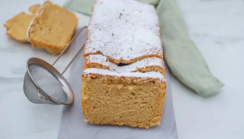 Puszyste ciasto keto, posypane cukrem pudrem, z dwoma kawałkami obok. Obok sitko do cukru.