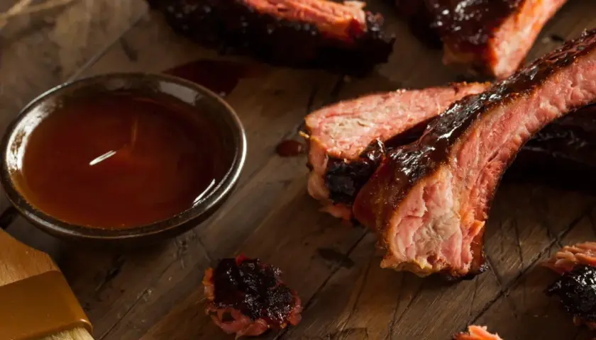 Soczyste żeberka BBQ, polane sosem, obok miseczka z marynatą do żeberek i pędzelek.