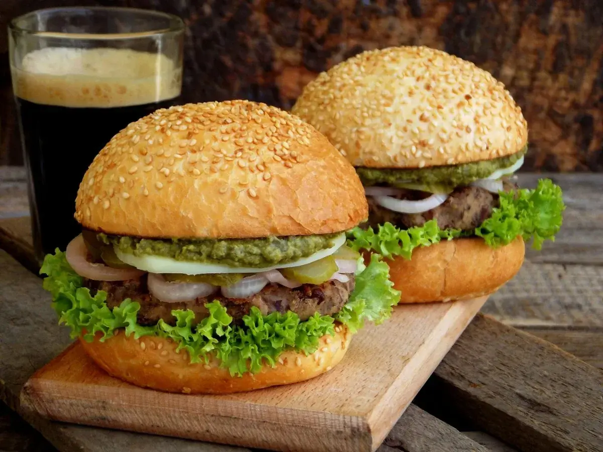Dwa domowe burgery z sezamem, sałatą, cebulą i sosem, obok szklanki ciemnego napoju.