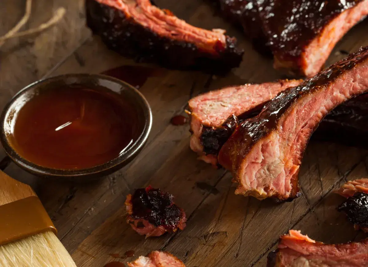 Soczyste żeberka BBQ, polane sosem, obok miseczka z marynatą do żeberek i pędzelek.