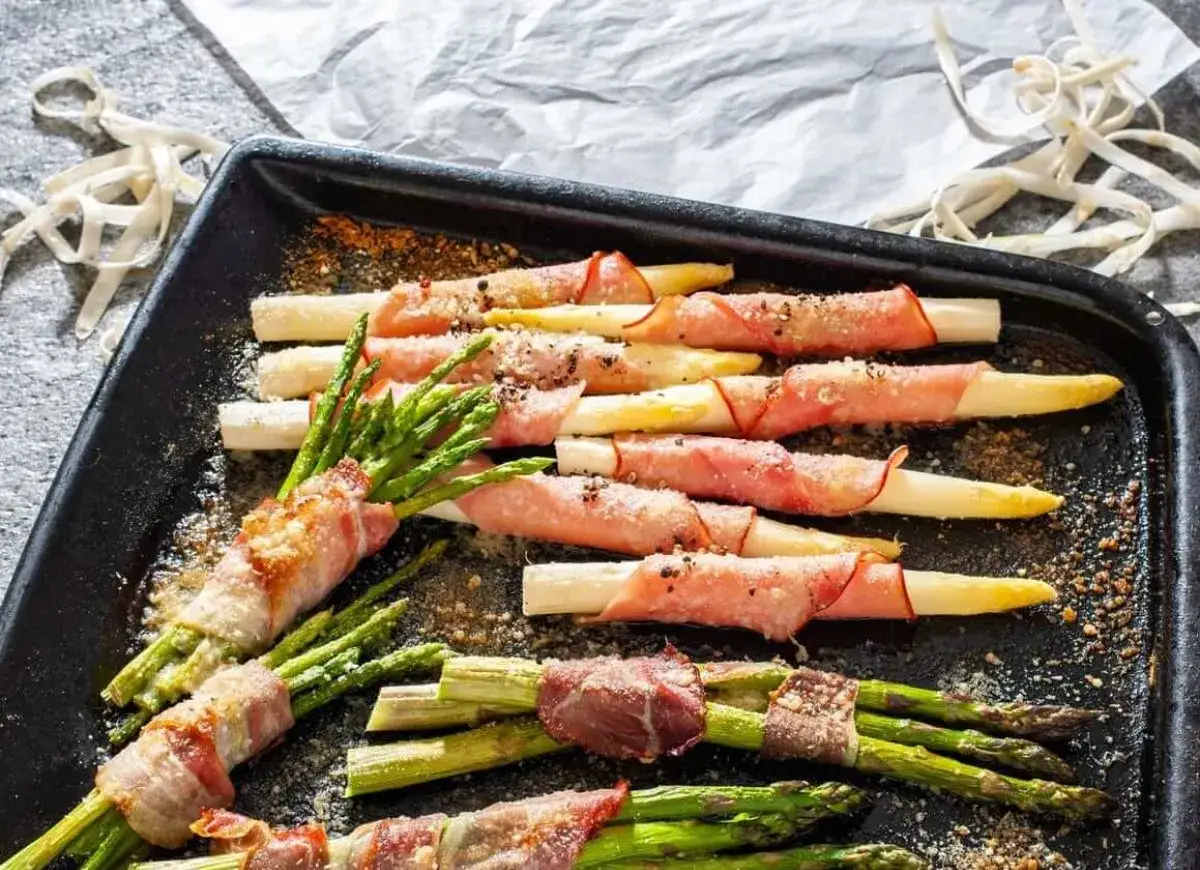 Zielone i białe szparagi owinięte szynką parmeńską, ułożone na czarnej blasze, posypane przyprawami.