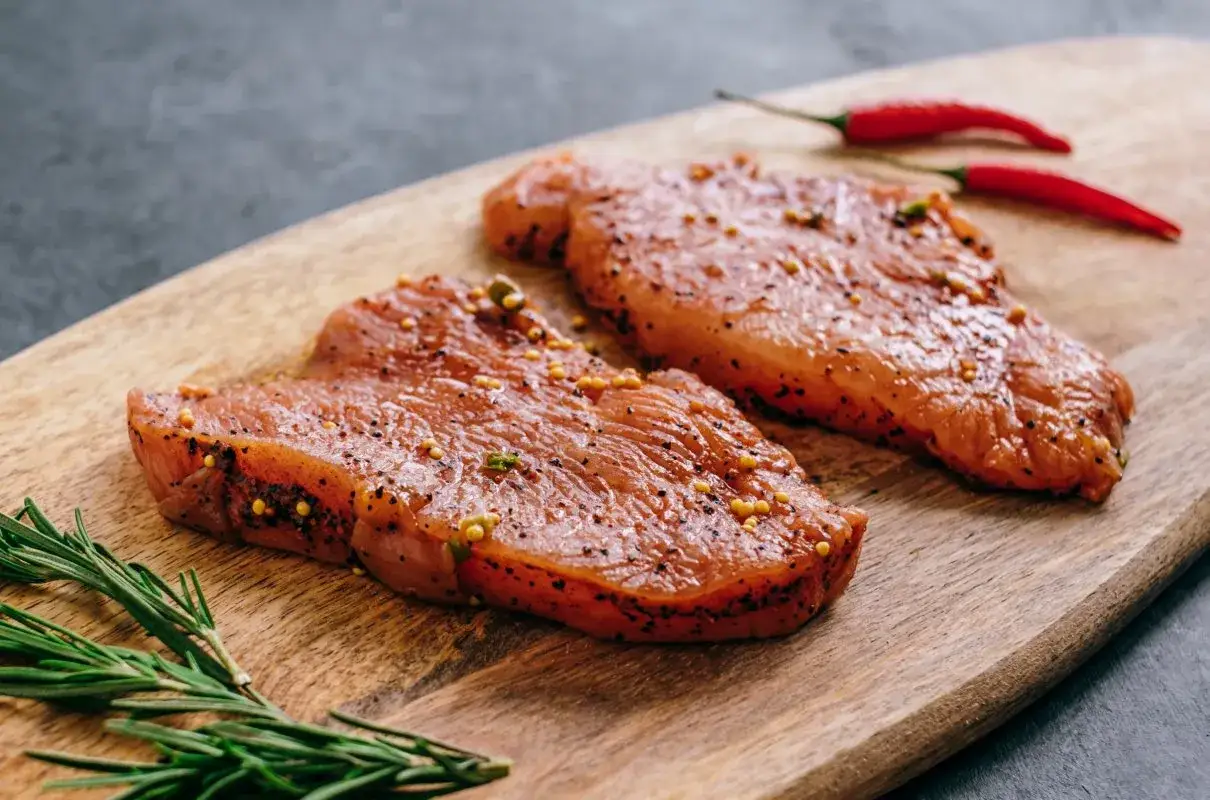 Dwa kawałki indyka w marynacie z pieprzem i musztardą, obok gałązka rozmarynu i dwa czerwone chili.