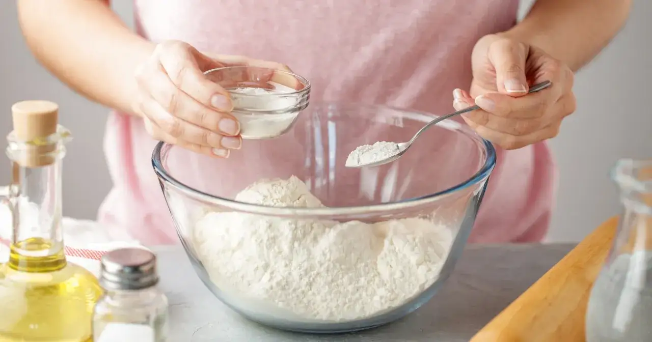 Ręce dodają sodę oczyszczoną do mąki w szklanej misce. Szukasz, czym zastąpić proszek do pieczenia? Soda to świetna alternatywa!